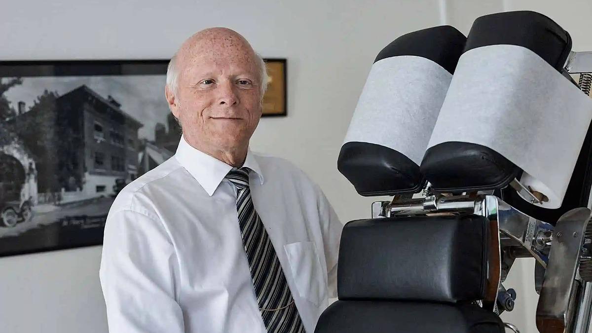 En ældre mand i skjorte og slips står ved en behandlingsstol i et klinikrum.