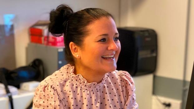En smilende kvinde med sort hår i en knold, iført en blomstret bluse, sidder ved en computer i et kontormiljø.
