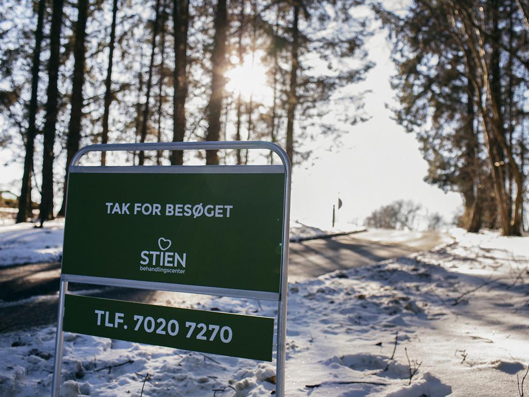Skilt med teksten "Tak for besøget" fra Stien, omgivet af sneklædte træer og en solstråle i baggrunden.