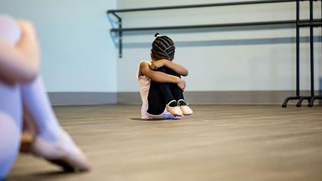 En lille pige sidder på gulvet i en dansestudio, hun krammer sine knæ og ser ned.