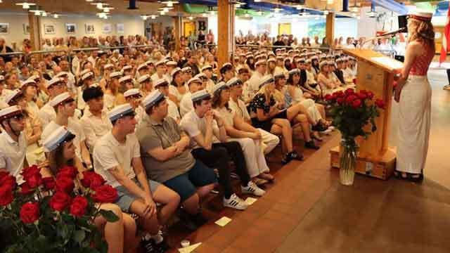En stor forsamling af studerende i hvide klæder og studenterhuer, der lytter til en taler ved en ceremoniel begivenhed.