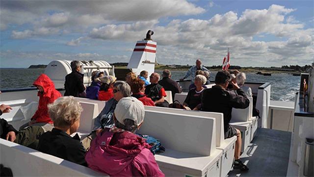Passagerer på en færge, der sejler over havet, med en klar himmel og grønne bakker i baggrunden.