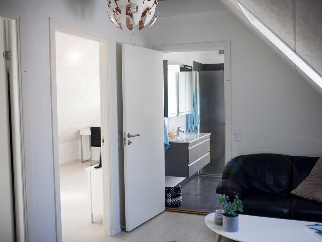 Stue med sort sofa, bord og åbne døre til badeværelse og kontor i en lys lejlighed.