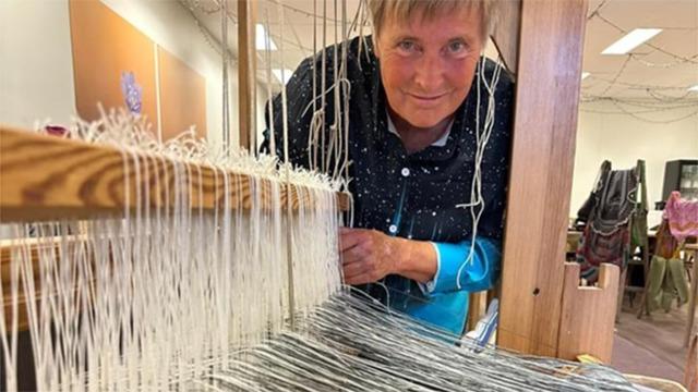 Kvindelig væver arbejder ved en væv med tråde i forskellige farver i et lyst rum.