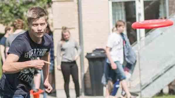 Ung mand kaster en frisbee på en udendørs legeplads, mens andre unge mennesker er i baggrunden.