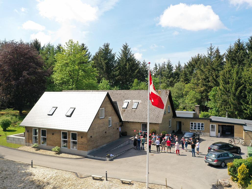 En gruppe mennesker samles foran et hus med en dansk flag i en skovklædt område.