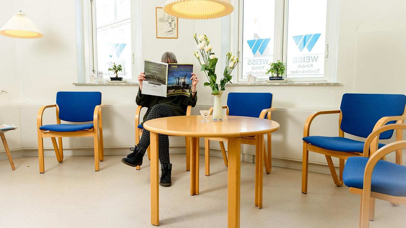 En person sidder i en venteværelse med blå stole og et rundt bord, mens de læser en avis.