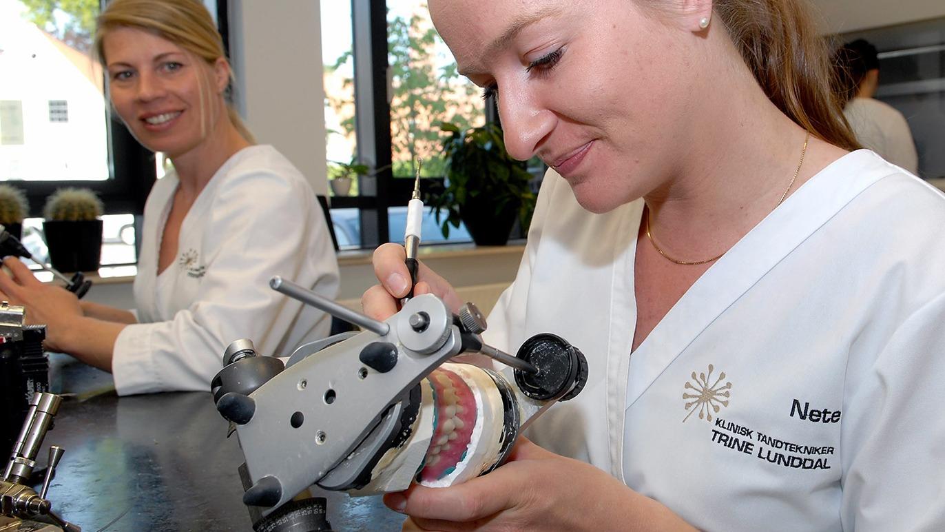 To kvinder arbejder med tandproteser i et laboratoriemiljø, hvor den ene kvinde finjusterer en tandprotes.