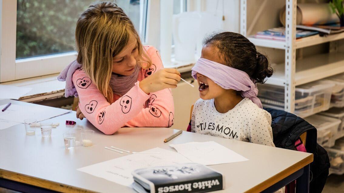 To piger leger en smagsprøveleg, hvor den ene er blindfoldet og får mad i munden.