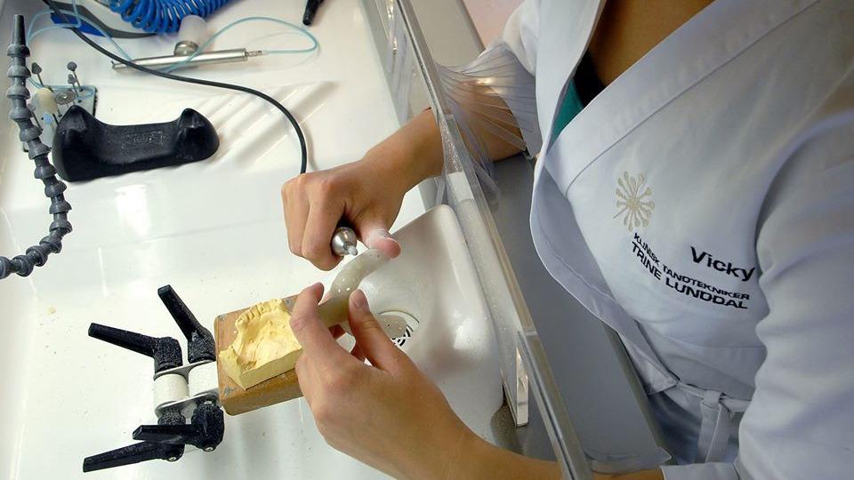 En person i hvid kittel arbejder med en tandprotes på et arbejdsbord med værktøj og udstyr.