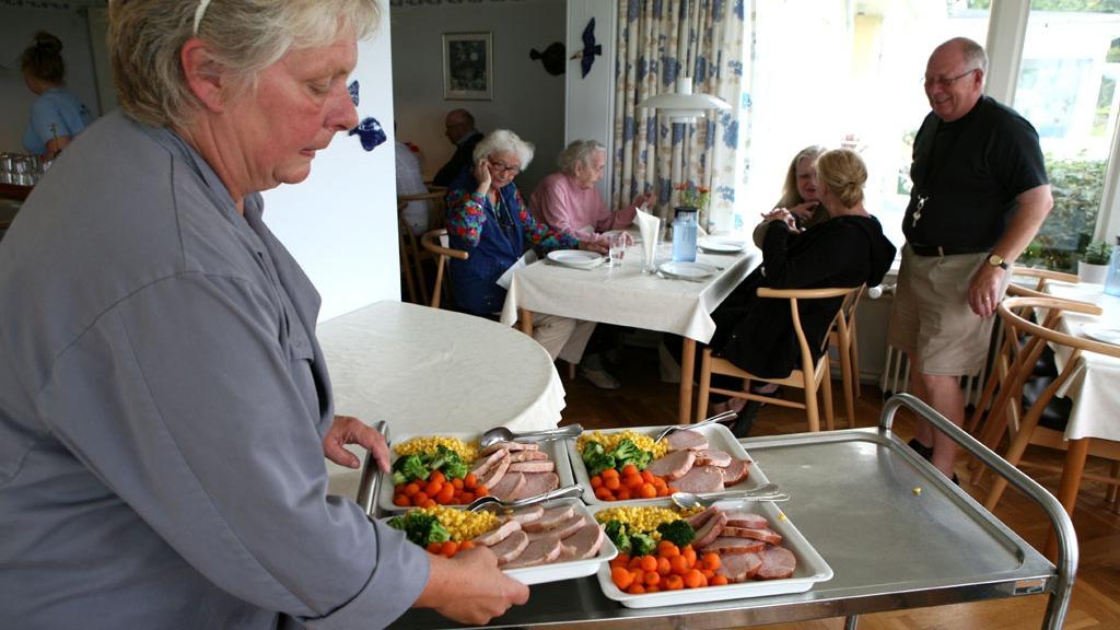En kvinde serverer mad på en vogn i en restaurant, mens gæsterne sidder ved bordene og snakker.