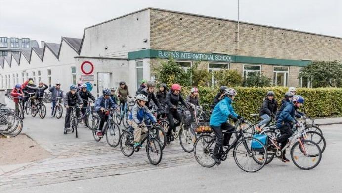 Børn og voksne cykler forbi en skole, iført hjelme og varme jakker, på en grå dag.