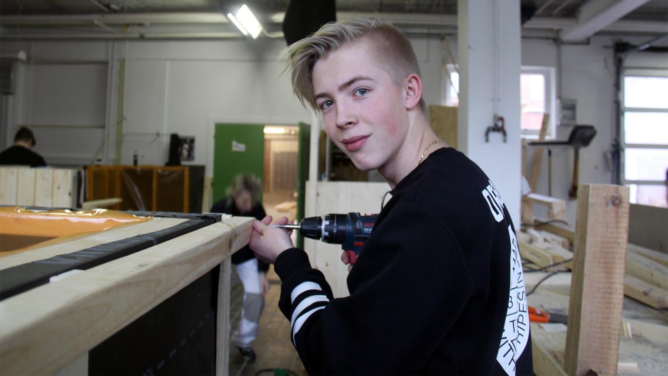 En ung dreng arbejder med en boremaskine i et værksted, omgivet af træmaterialer og andre elever.