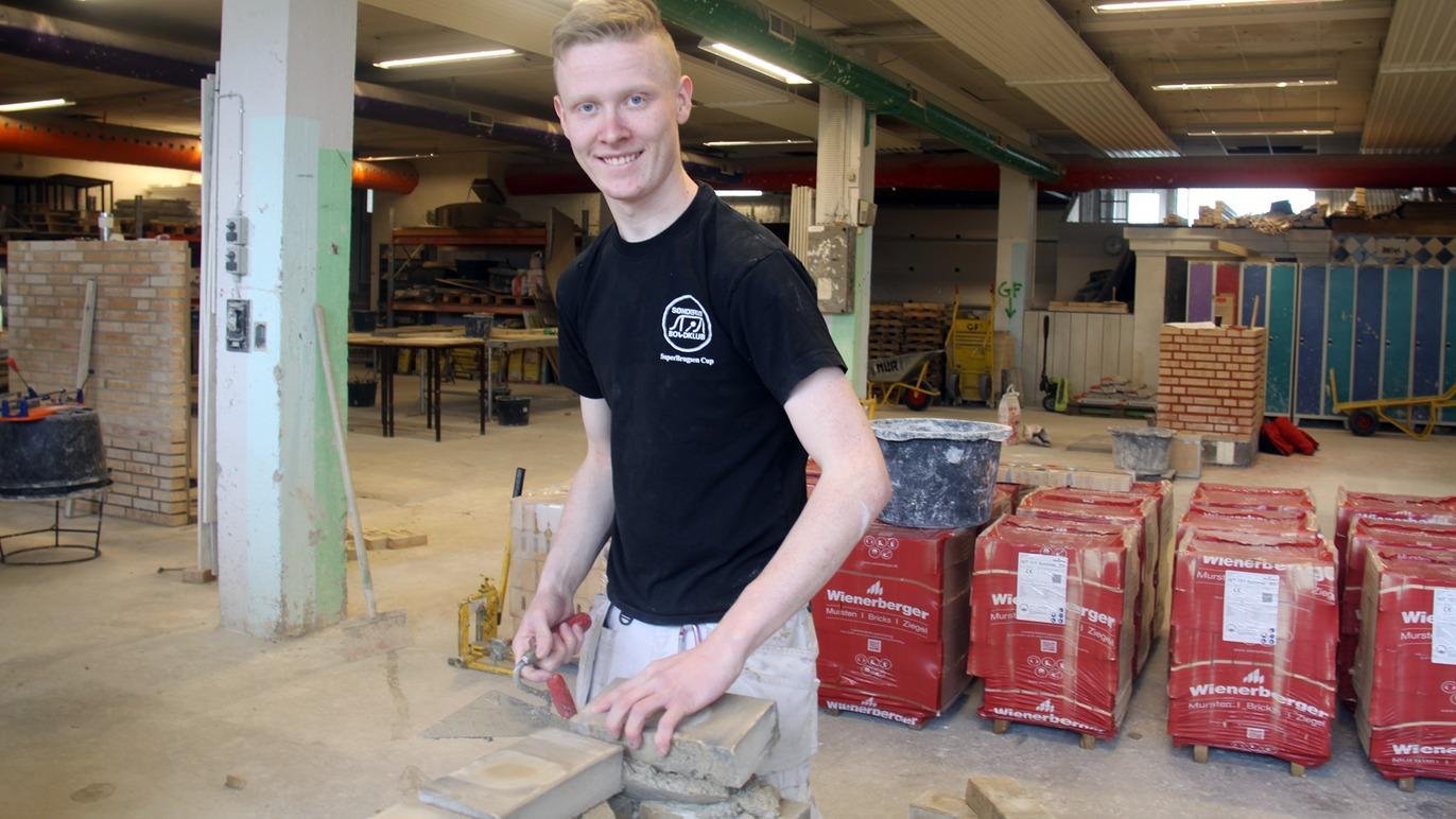 En smilende ung mand arbejder med mursten i et byggeværksted, omgivet af materialer og værktøj.