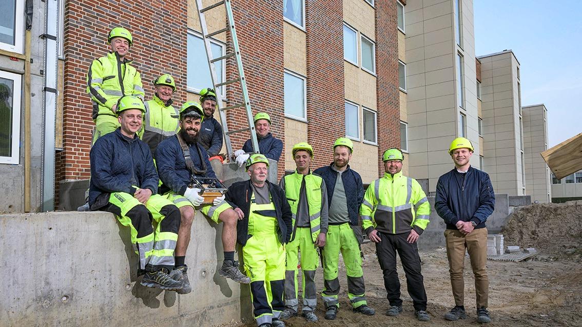 Gruppe af bygningsarbejdere i sikkerhedshjelme og arbejdstøj foran en bygning under konstruktion.