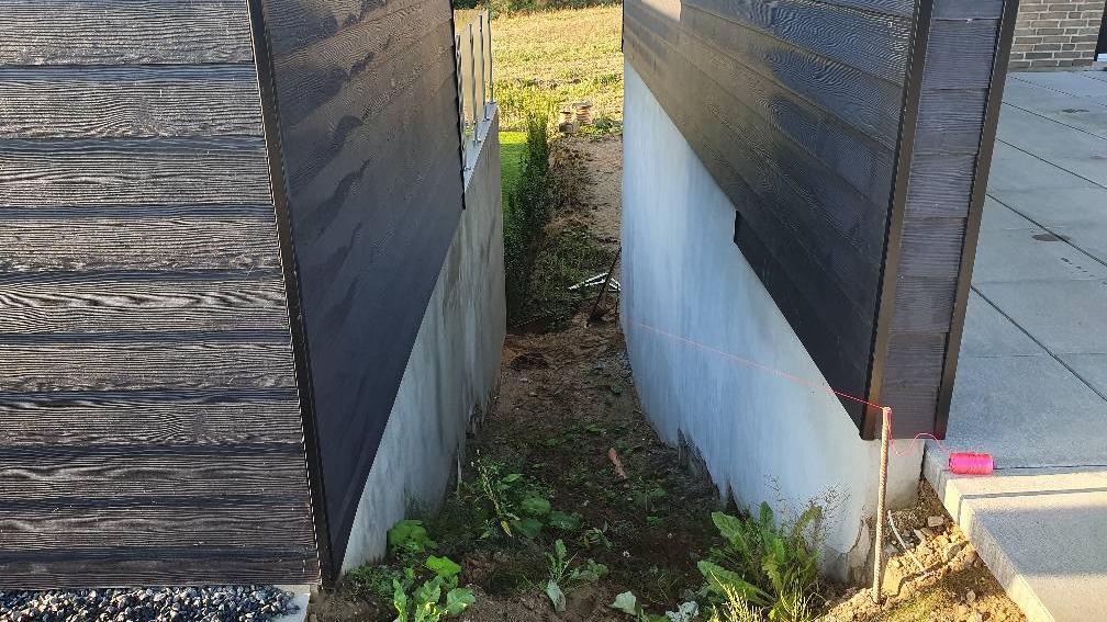 Smal passage mellem to bygninger med jord og planter i bunden.