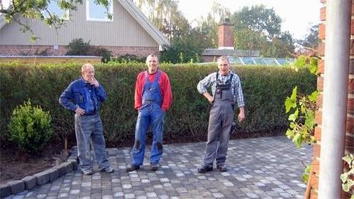 Tre mænd i arbejdstøj står på en nybelagt flisebelægning foran en hæk og et hus.