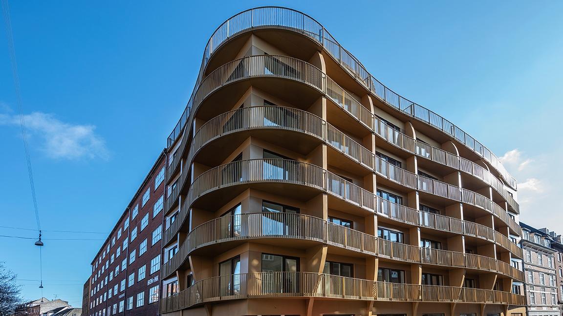 Moderne bygning med buet facade og altaner, beliggende under en klar blå himmel.