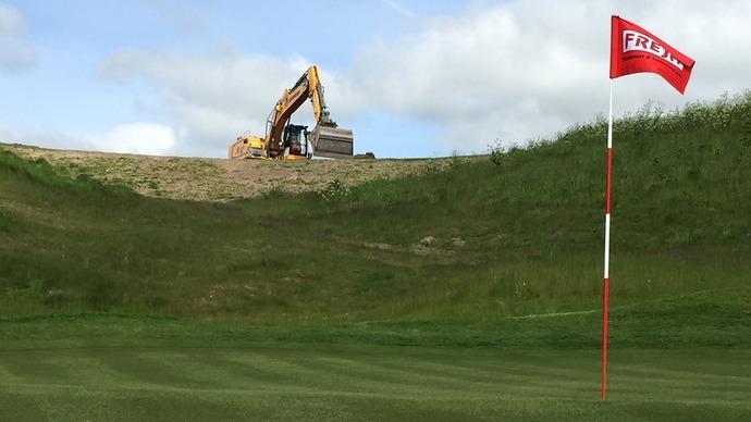 Grøn golfbane med en flagstang i forgrunden og en gravemaskine, der arbejder på en bakke i baggrunden.