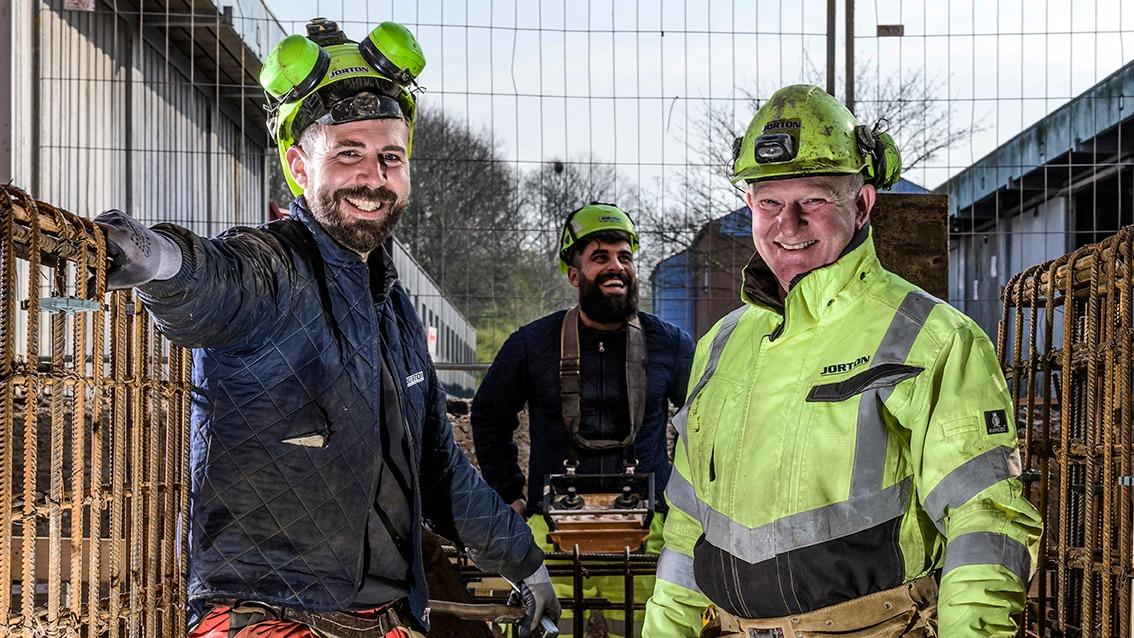 Tre glade arbejdere i sikkerhedsudstyr står foran et byggeområde med hegn.