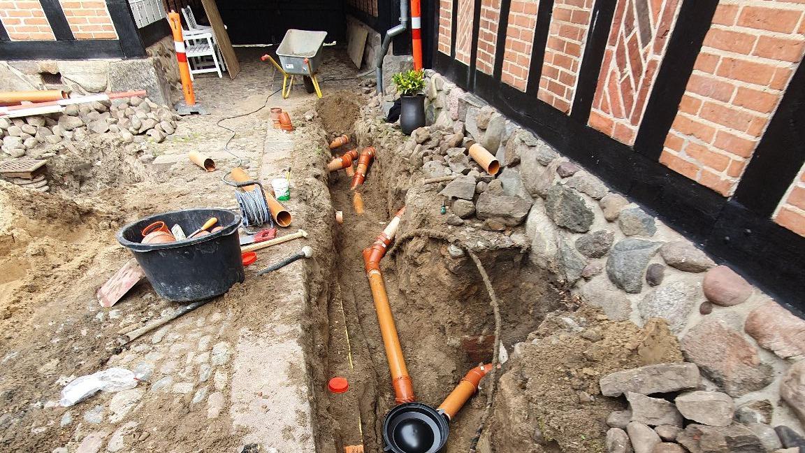 Gravested med rørinstallation, grus og værktøj ved en bygning med bindingsværk.