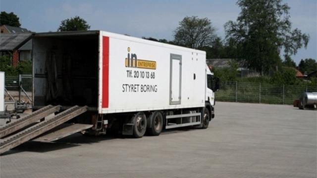 En lastbil med en rampe parkeret på en grusplads, med teksten "STYRET BOLIG" på siden.