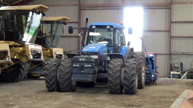 En blå traktor står i en lade med landbrugsmaskiner og sollys, der strømmer ind gennem vinduet.