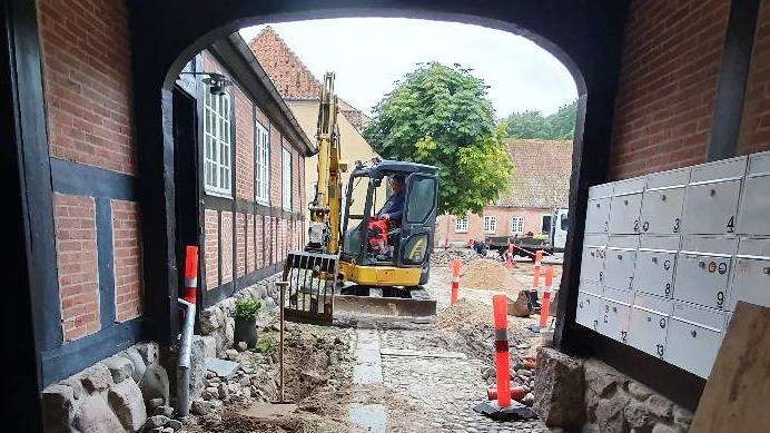 En gravemaskine arbejder på en byggeplads med røde afspærringer og en bygning i baggrunden.