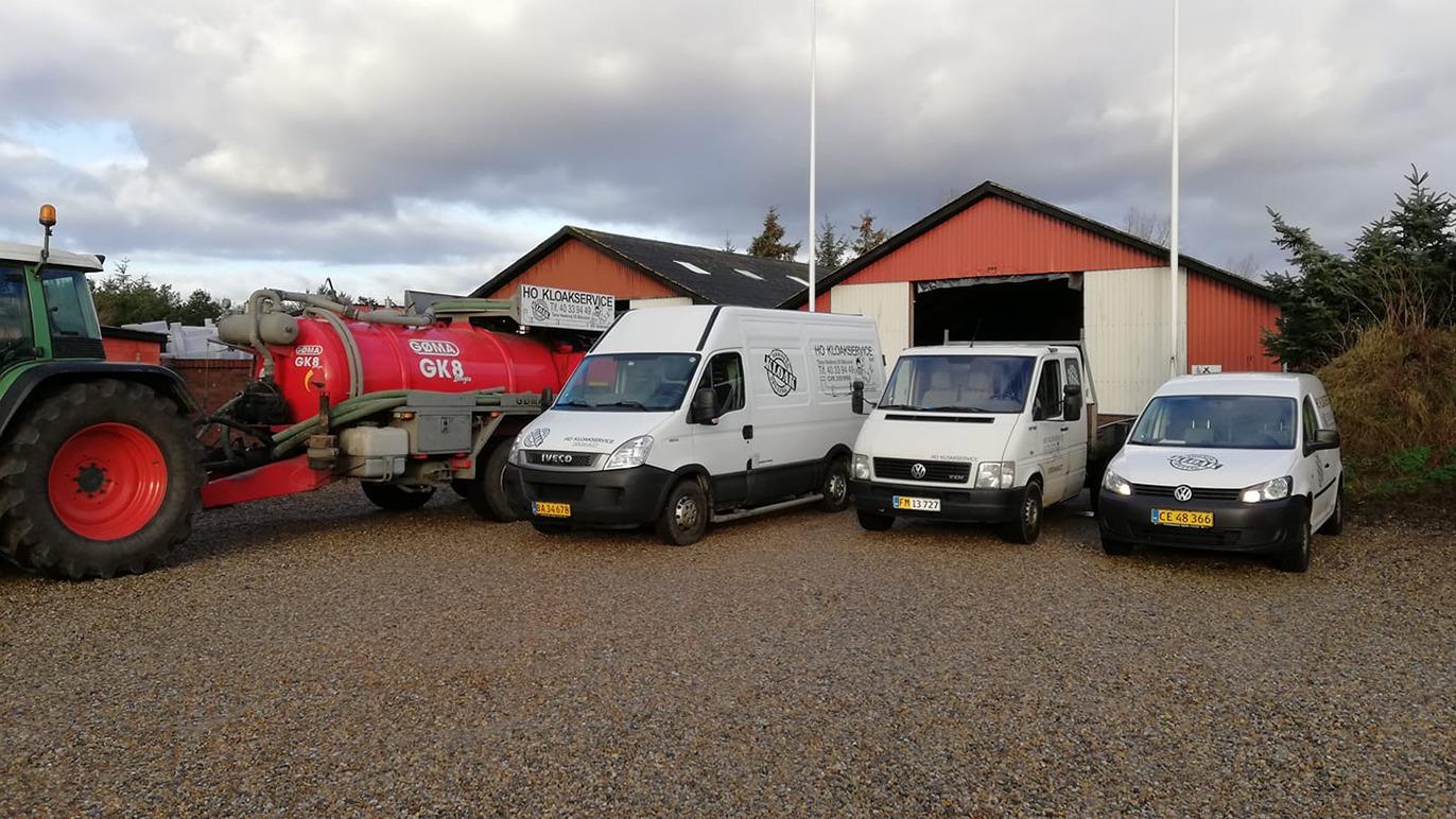 Tre hvide varevogne og en traktor parkeret foran en rød lade med grusbelægning.