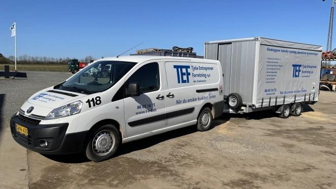 En hvid varevogn med trailer, der har TEF-logoet, parkeret på en grusplads under en blå himmel.