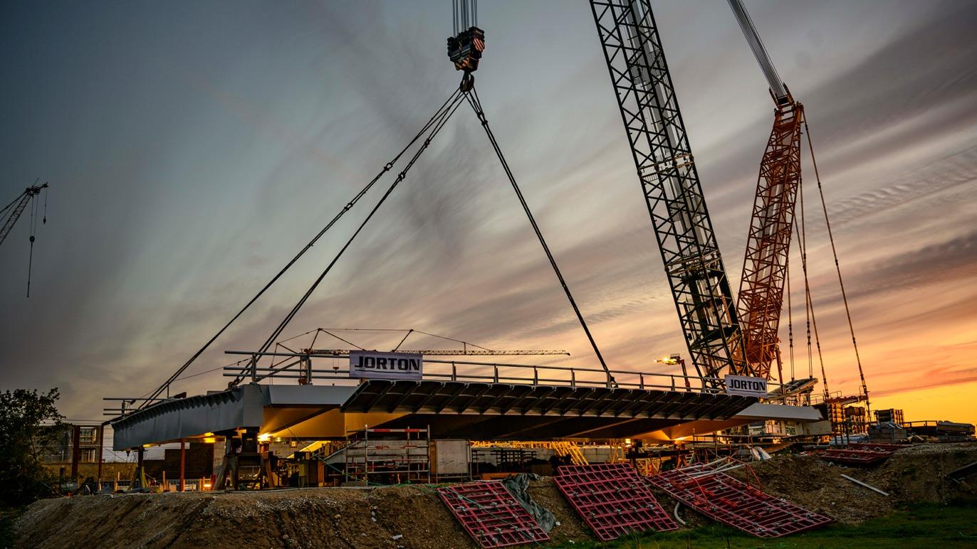 Byggeri af en bro med kraner, der løfter en betonplade ved solnedgang.