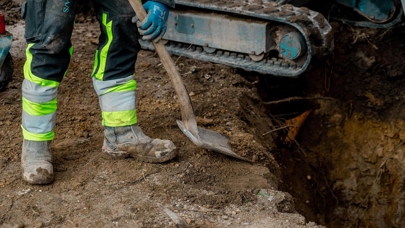 En arbejdstager graver i jorden med en skovl ved en gravemaskine på en byggeplads.
