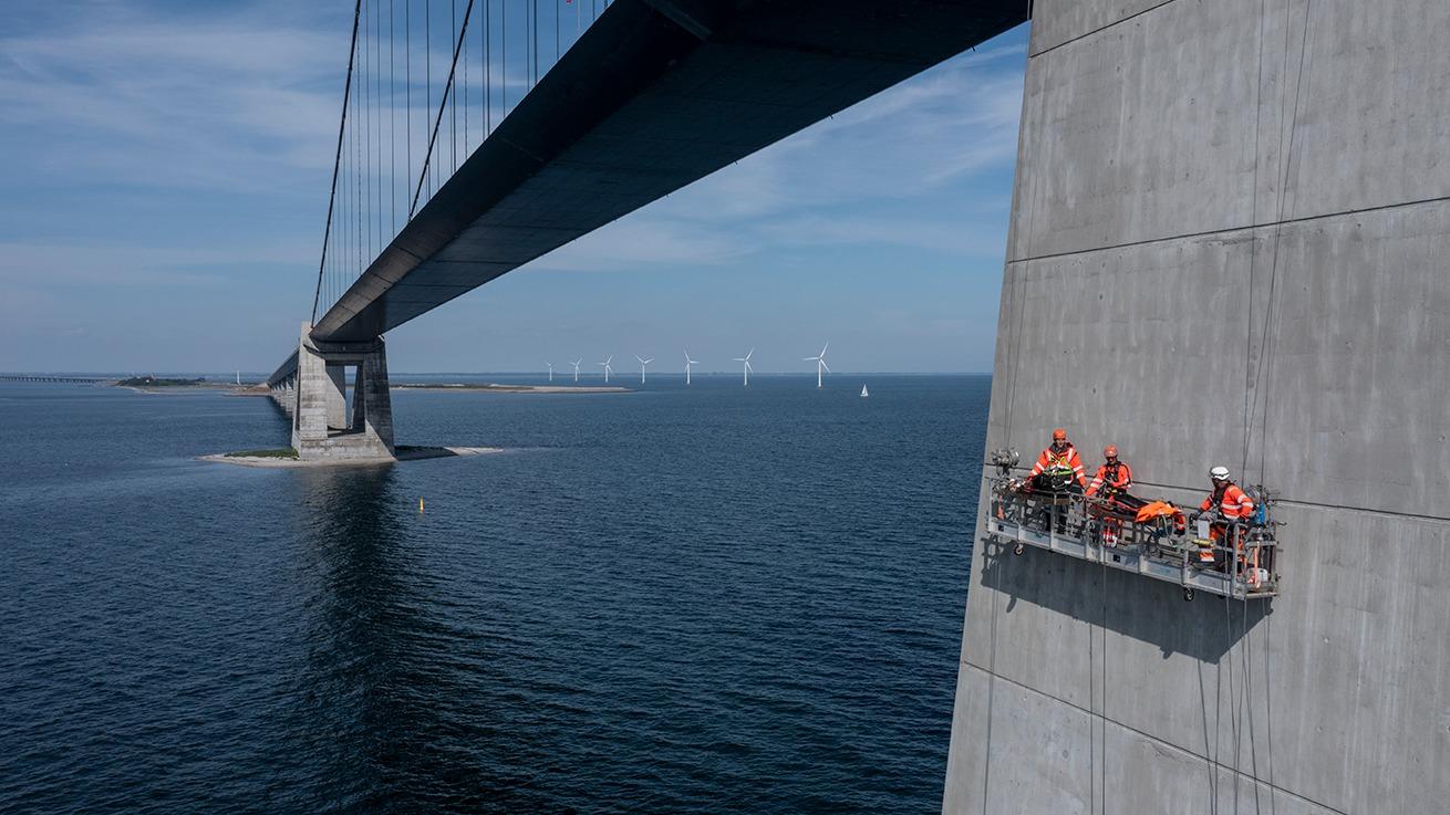 Arbejdsfolk på en platform under en bro, med vand og vindmøller i baggrunden.