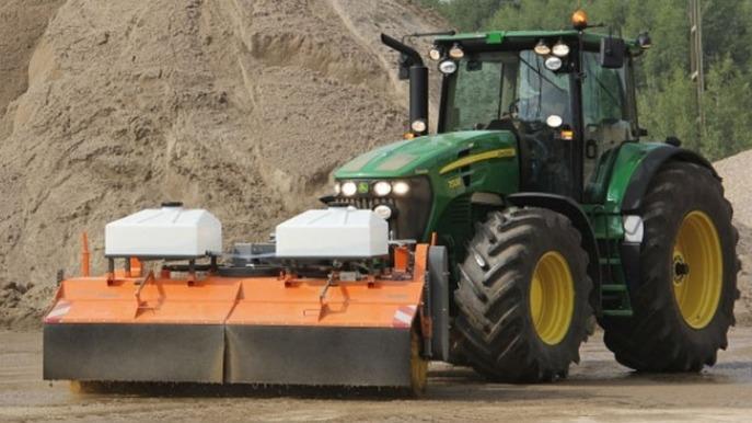 En grøn traktor med en orange sneslynge arbejder på en byggeplads med sand og grus.