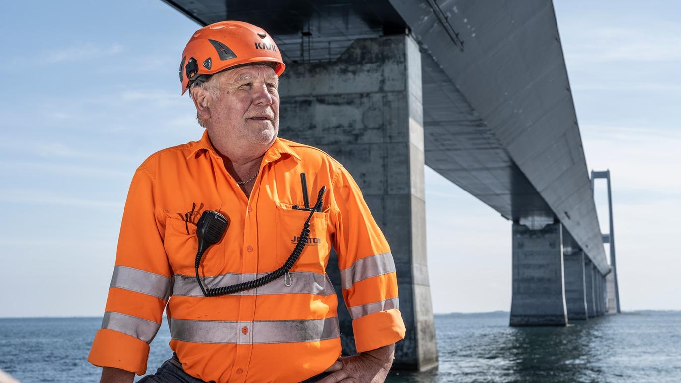 En mand i orange sikkerhedstøj og hjelm står under en bro ved vandet.