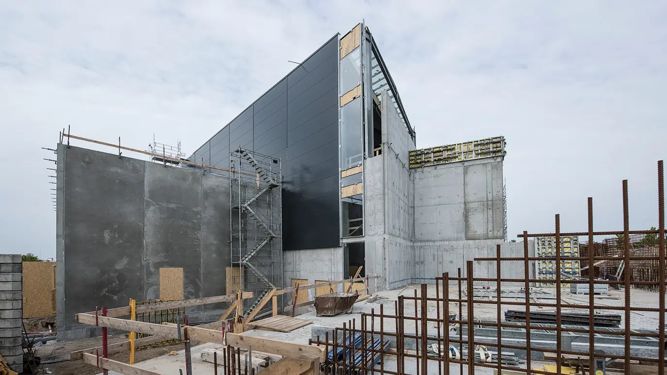 Byggeri af moderne bygning med betonvægge og metalfacade under konstruktion. Stålrammer og stilladser er synlige.