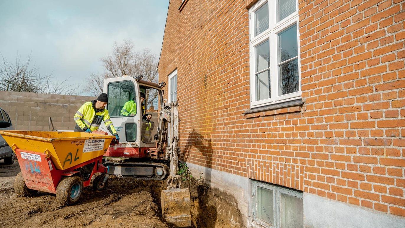 En gravemaskine arbejder ved en murstensvæg, mens en arbejder i sikkerhedstøj overvåger processen.