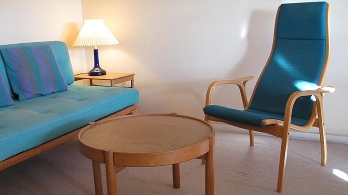 Stue med en blå sofa, en høj stol og et rundt bord, oplyst af en lampe.