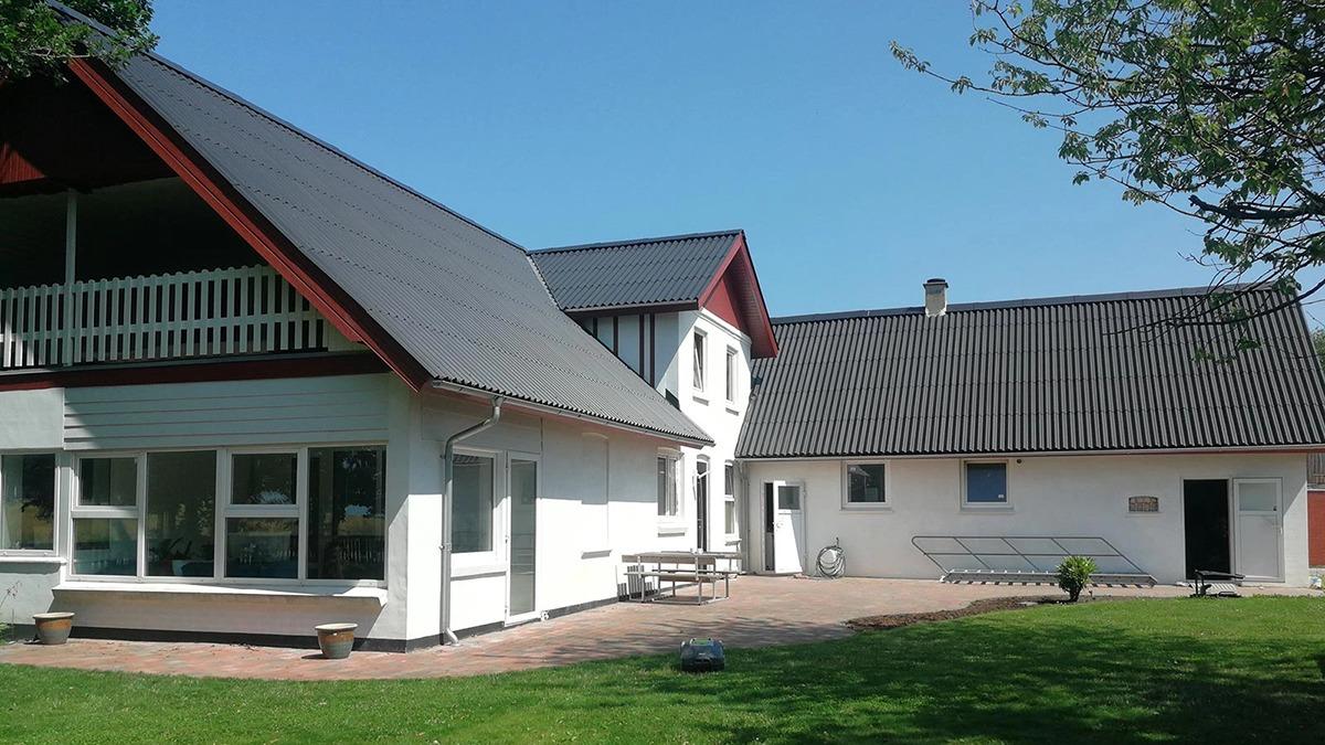 Et moderne hus med to etager og en terrasse, omgivet af grønne træer og en klar blå himmel.
