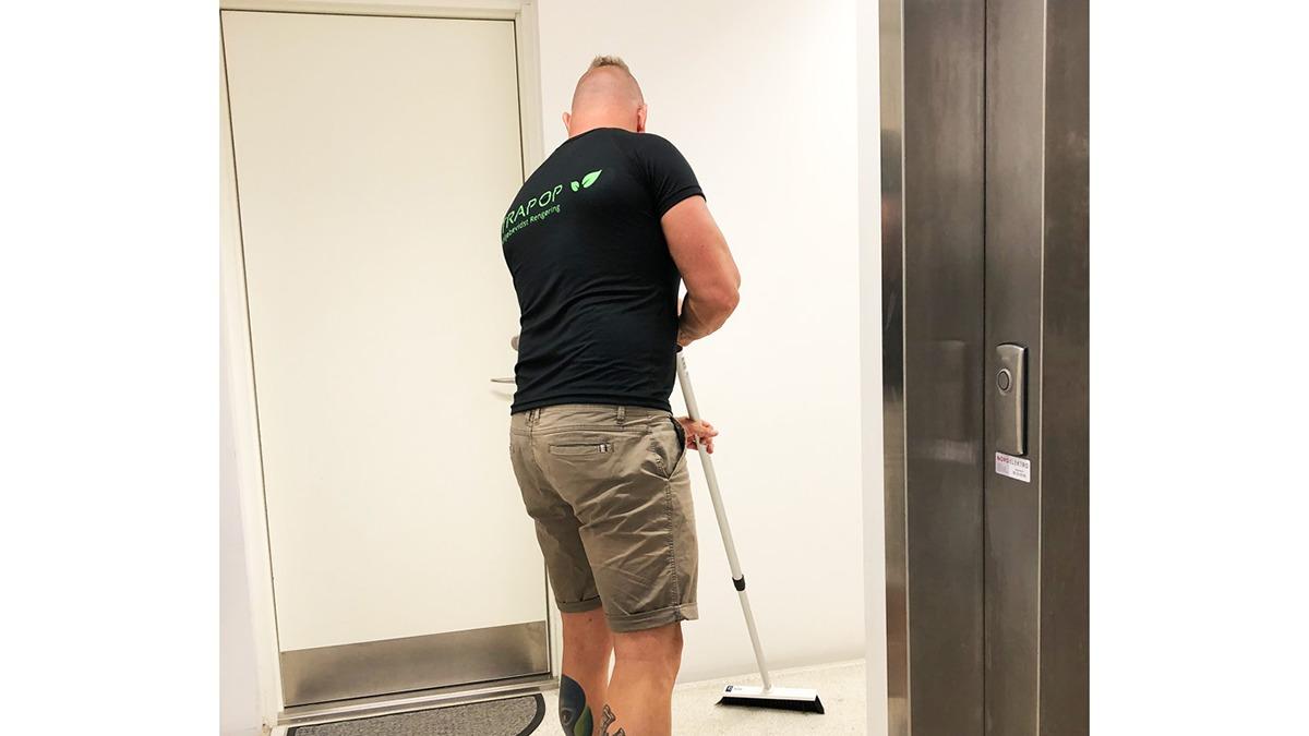 Mand der støvsuger en gang i en bygning, iført sort t-shirt og shorts, med en dør og elevator i baggrunden.