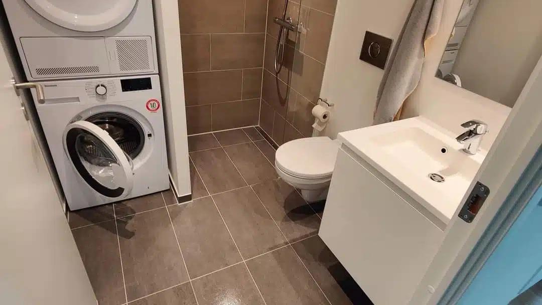 Badeværelse med vaskemaskine, tørretumbler, toilet og moderne håndvask med skab. Fliser på gulvet og væggene.