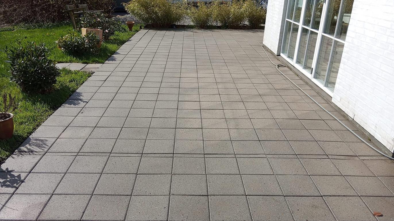 Flisebelagt terrasse med grønne planter og buske i baggrunden. Solskin på fliserne.