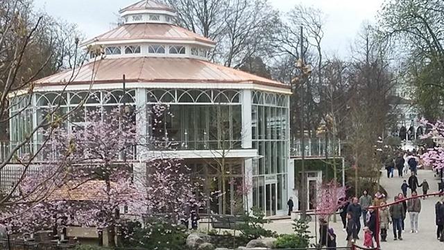 En smuk pavillon omgivet af blomstrende kirsebærtræer i en park med besøgende, der nyder det gode vejr.