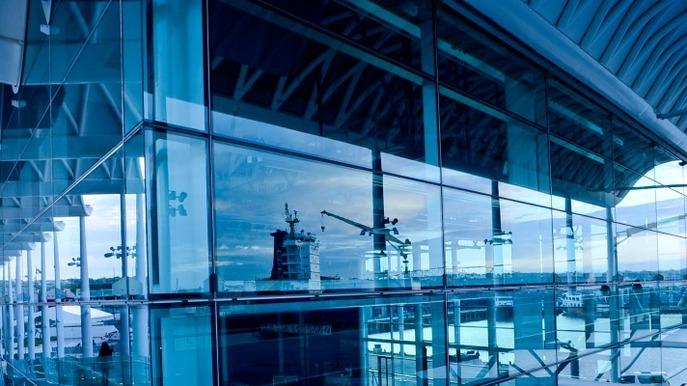 Moderne lufthavnsterminal med glasvægge og refleksioner af kraner og himmel i blå nuancer.