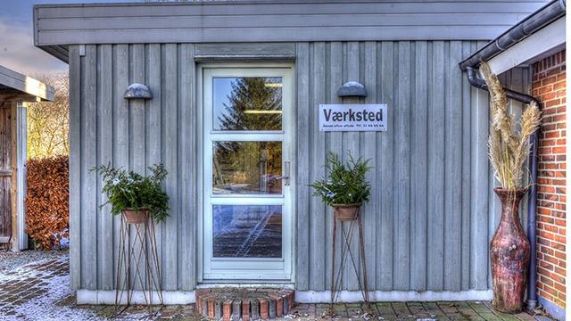 Værksted med grå træfacade, hvid dør og planter i krukker ved indgangen.