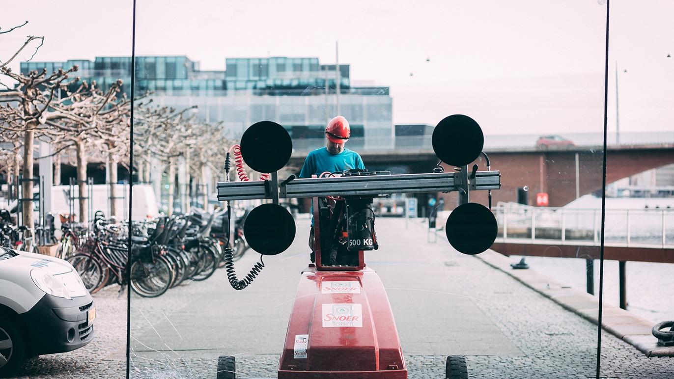 En person i rød hjelm kører en stor maskine med vægte langs en cykelsti ved vandet.