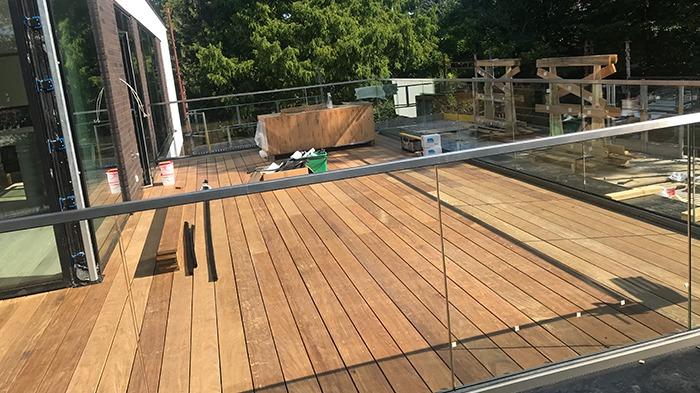 Ny terrasse med træplanker og glasrækværk, omgivet af grønne træer og byggeudstyr.