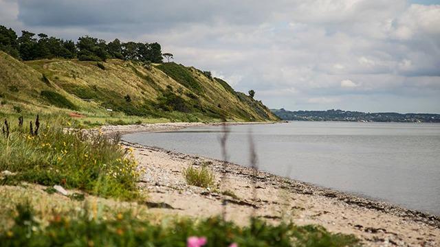 En smuk kystlinje med grønne skråninger og stille vand under en skyet himmel.