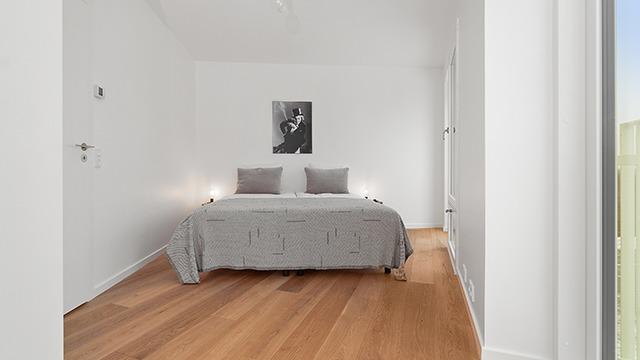 Soveværelse med dobbeltseng, grå sengetæppe, to puder og moderne kunst på væggen. Trægulv og lys indretning.