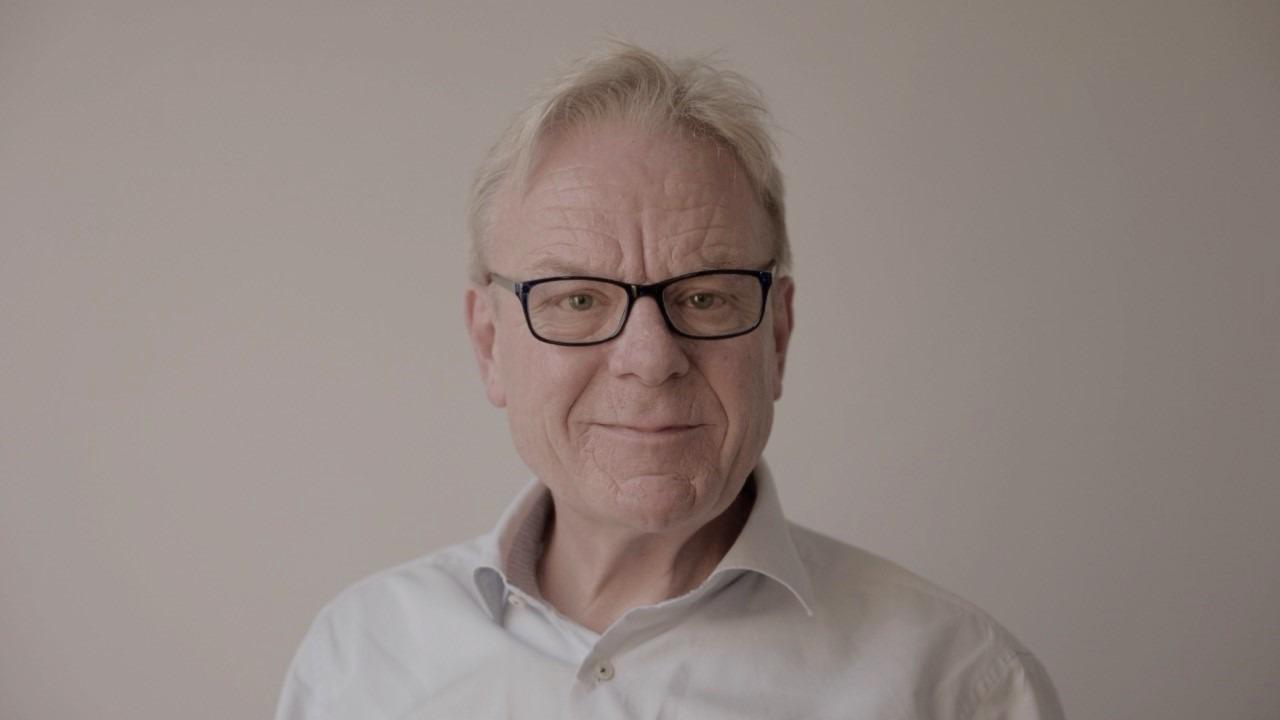 En ældre mand med briller og gråt hår, iført en hvid skjorte, står foran en lys baggrund.